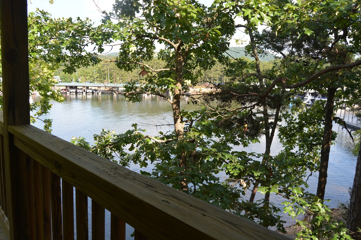 A view of the lake is framed by lush green trees from a wooden deck. The water reflects the surrounding landscape, with boats visible in the distance, enhancing the serene lakeside atmosphere.