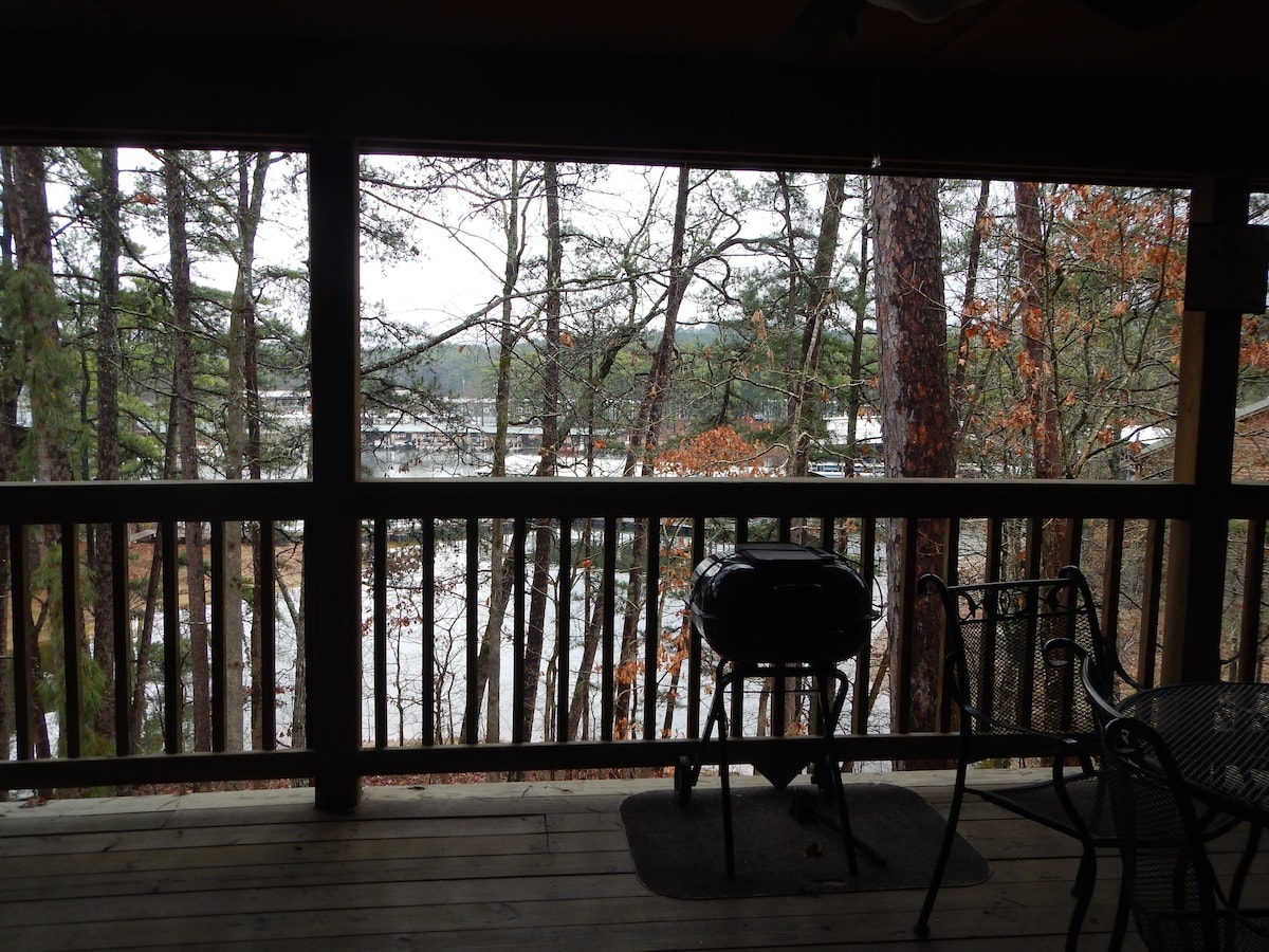 A spacious deck overlooks a serene view of the marina, framed by tall trees. A charcoal grill is positioned beside a set of black metal chairs and table, inviting outdoor dining amid nature.