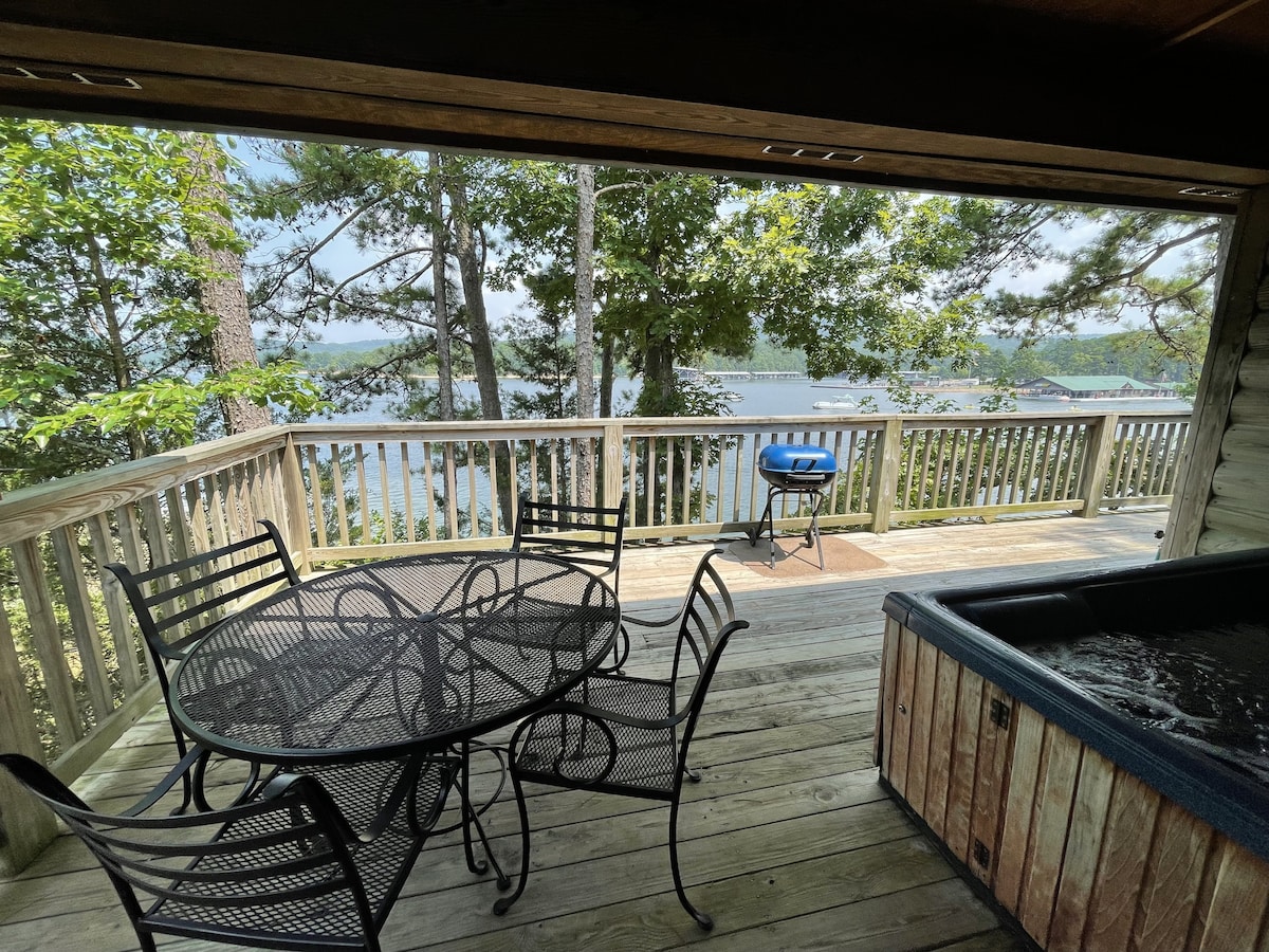 A spacious wooden deck is presented, featuring a round table surrounded by four metal chairs. Nearby, a hot tub is positioned next to a charcoal grill, with a scenic view of the lake visible through trees in the background.