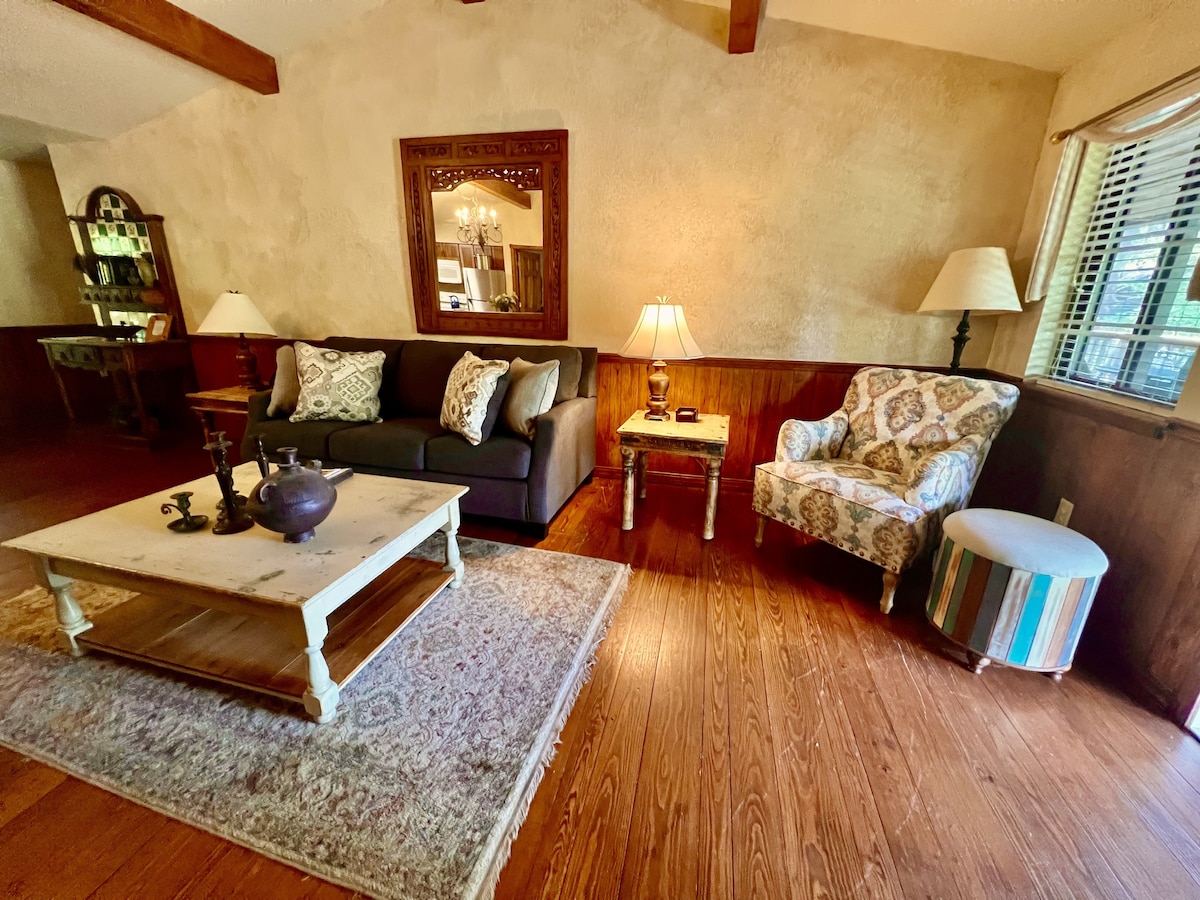 A spacious living area features a dark sofa paired with an ornate armchair and a striped ottoman. A large coffee table sits on a decorative rug, illuminated by soft lighting from table lamps. Natural light enters through the window, accentuating the warm wooden floors.