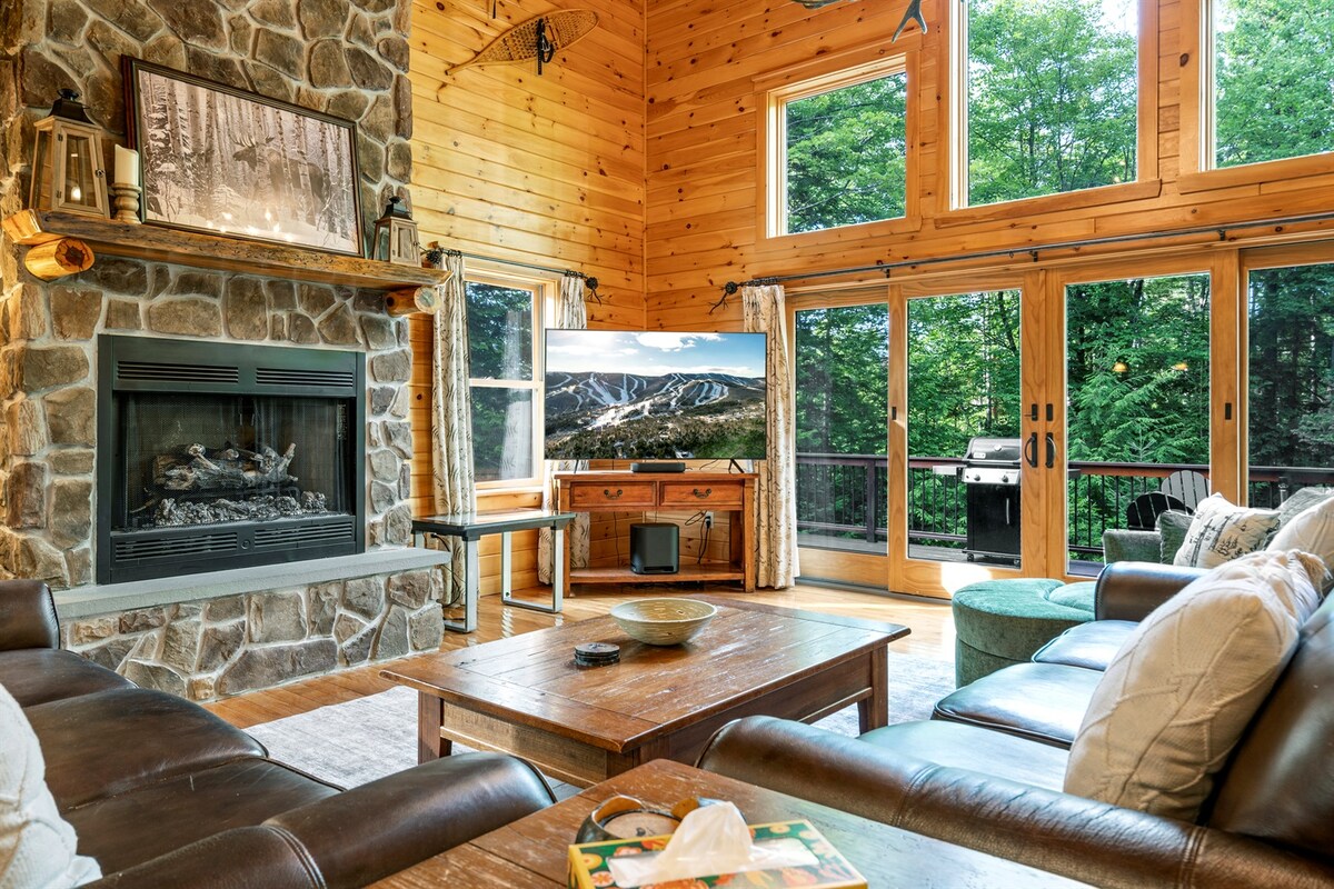 A cozy living area features a stone fireplace surrounded by wood paneling, enhancing a rustic charm. Large windows frame views of the trees outside, while a flat-screen TV is positioned atop a wooden console. Comfortable seating surrounds a center coffee table.