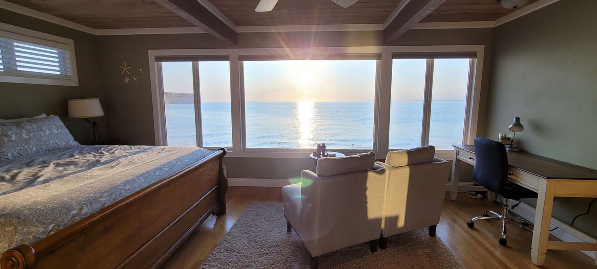 A spacious primary bedroom features a king bed positioned to overlook the ocean through large windows, allowing natural light to flood the space. A cozy seating area with cushioned chairs is present, along with a work desk, creating an inviting atmosphere to enjoy the serene views.