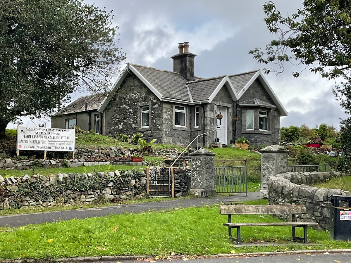 A stone cottage is set amidst a green landscape, featuring a pitched roof and large windows. A well-maintained garden surrounds the property, with trees and shrubs enhancing the natural setting. A bench is positioned nearby, offering a space to relax and enjoy the view.
