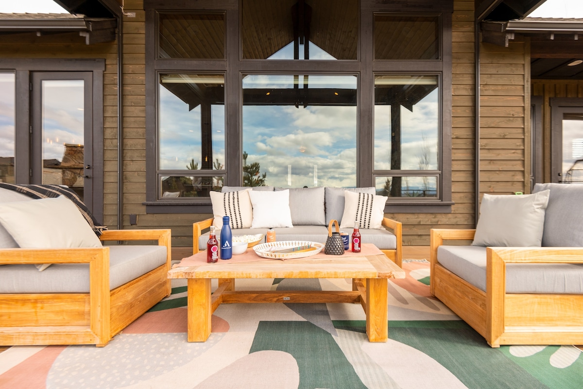 An outdoor lounge area is furnished with wooden seating and plush cushions, situated on a patterned rug. A coffee table is arranged with decorative items and drinks, while large windows reflect the surrounding landscape and provide ample natural light.