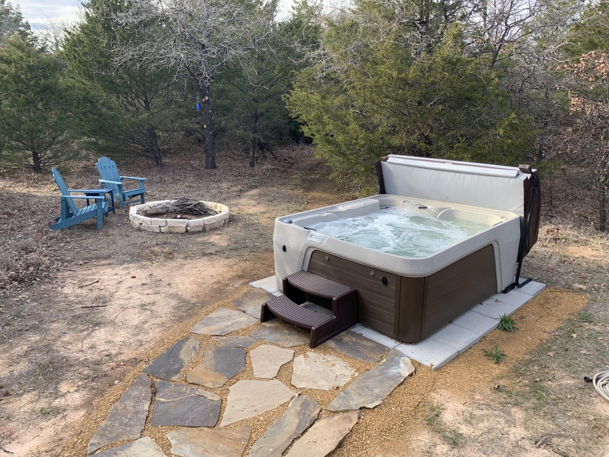 A hot tub is positioned beside a stone pathway, leading to a substantial fire pit surrounded by seating. The area is bordered by trees, creating a secluded atmosphere. Natural ground and stones complete the tranquil outdoor space.