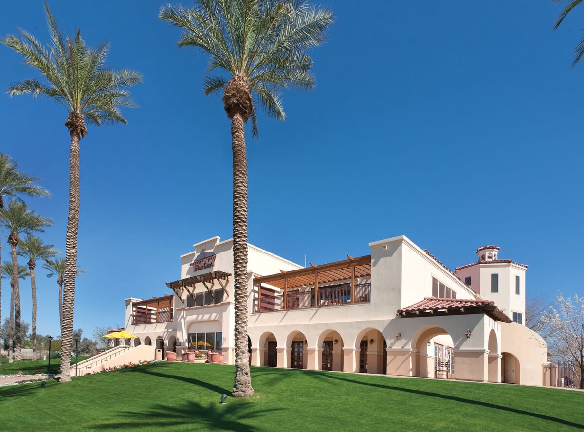The resort's exterior is showcased against a clear blue sky, featuring a spacious lawn bordered by tall palm trees. The building incorporates an arched design with a shaded patio area, providing a welcoming space for guests to relax.