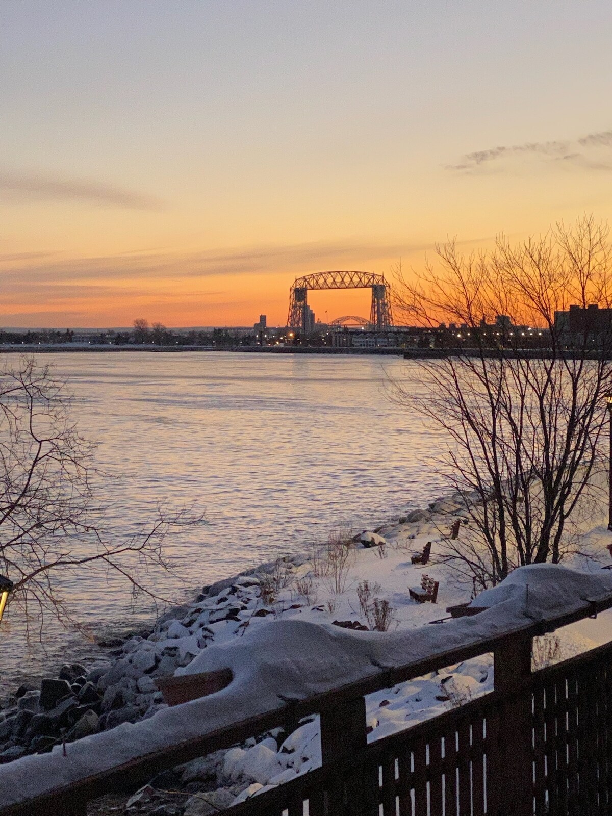 A scenic view of Lake Superior at sunset, with soft hues of orange and pink reflected on the water. The iconic Duluth Aerial Lift Bridge is visible in the distance, framed by snow-covered rocks and bare trees along the shoreline.
