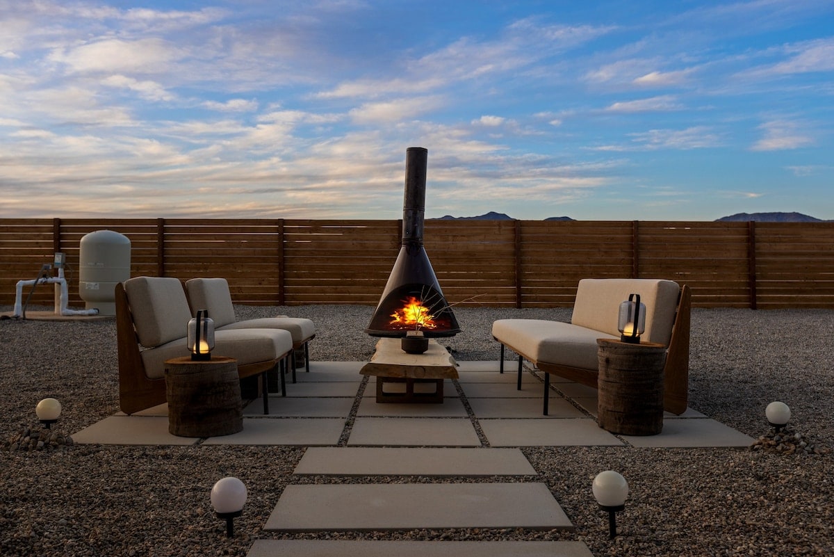 A modern outdoor seating area is set on a gravel surface, featuring a fire pit at the center. Two comfortable benches and a low table are arranged around the fire, with lanterns providing soft illumination. The backdrop includes a wooden fence and expansive sky.