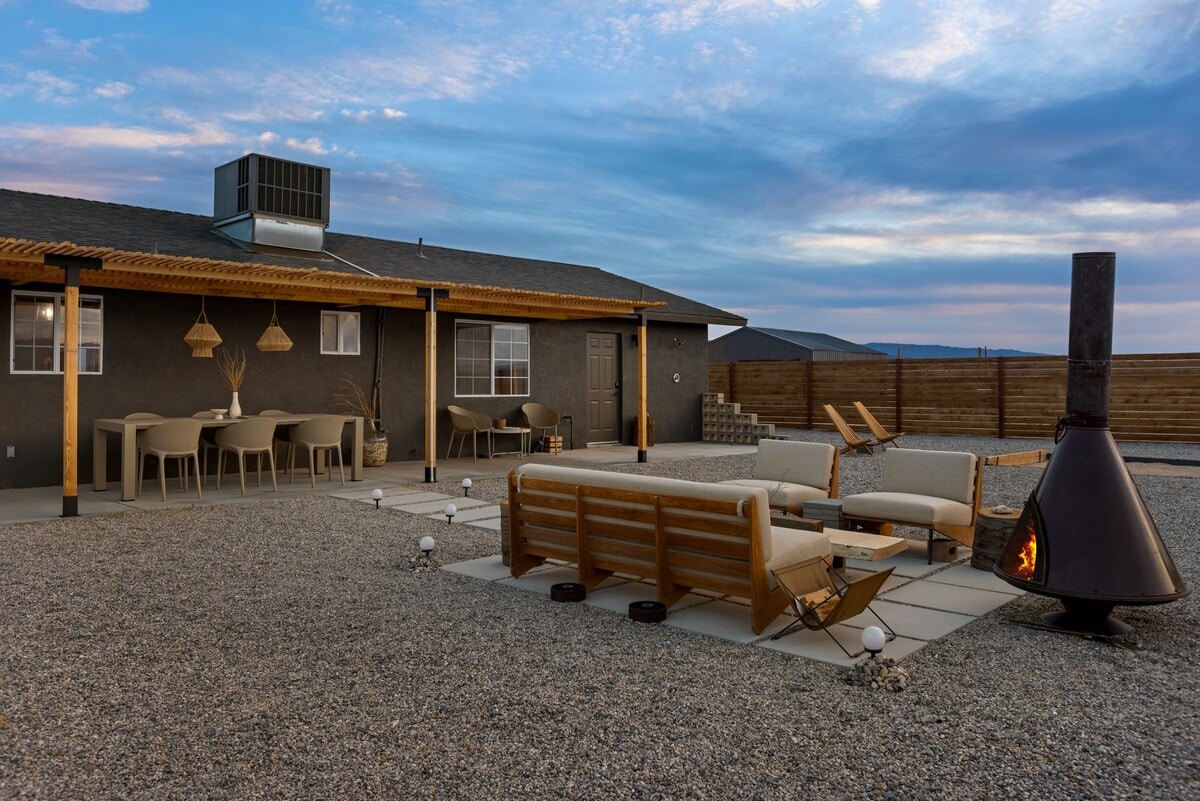 An outdoor living area is featured, showcasing a cozy seating arrangement with wooden benches and cushions. A vintage firepit sits nearby, with an inviting glow. A spacious dining table is positioned under a covered patio, surrounded by gravel, and distant views of the expansive desert landscape.