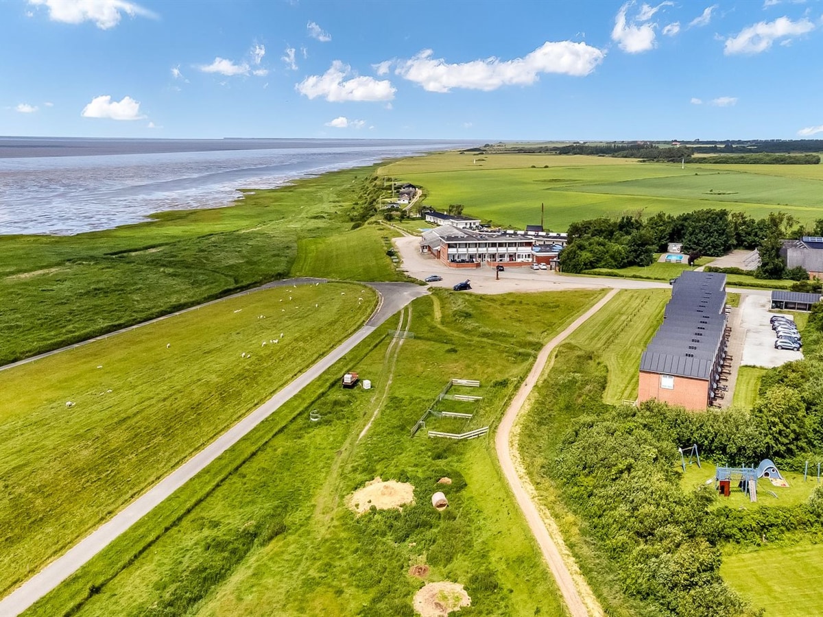 The image captures a scenic aerial view of the surrounding landscape, featuring expansive green fields and a meandering pathway. The coastline is visible in the distance, with a gentle waterway on the right. A small collection of buildings can be seen near the road that curves through the area.