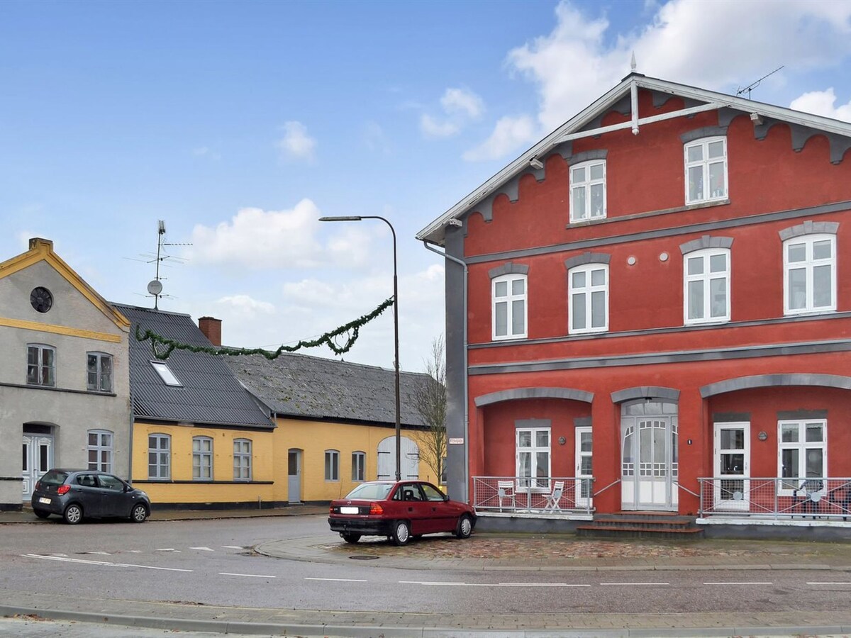 A vibrant red multi-story building is visible alongside a yellow structure and a gray building, set at a street corner. The architecture reflects a variety of styles. Cars are parked nearby, and greenery is positioned along the street. The sky is bright with scattered clouds.