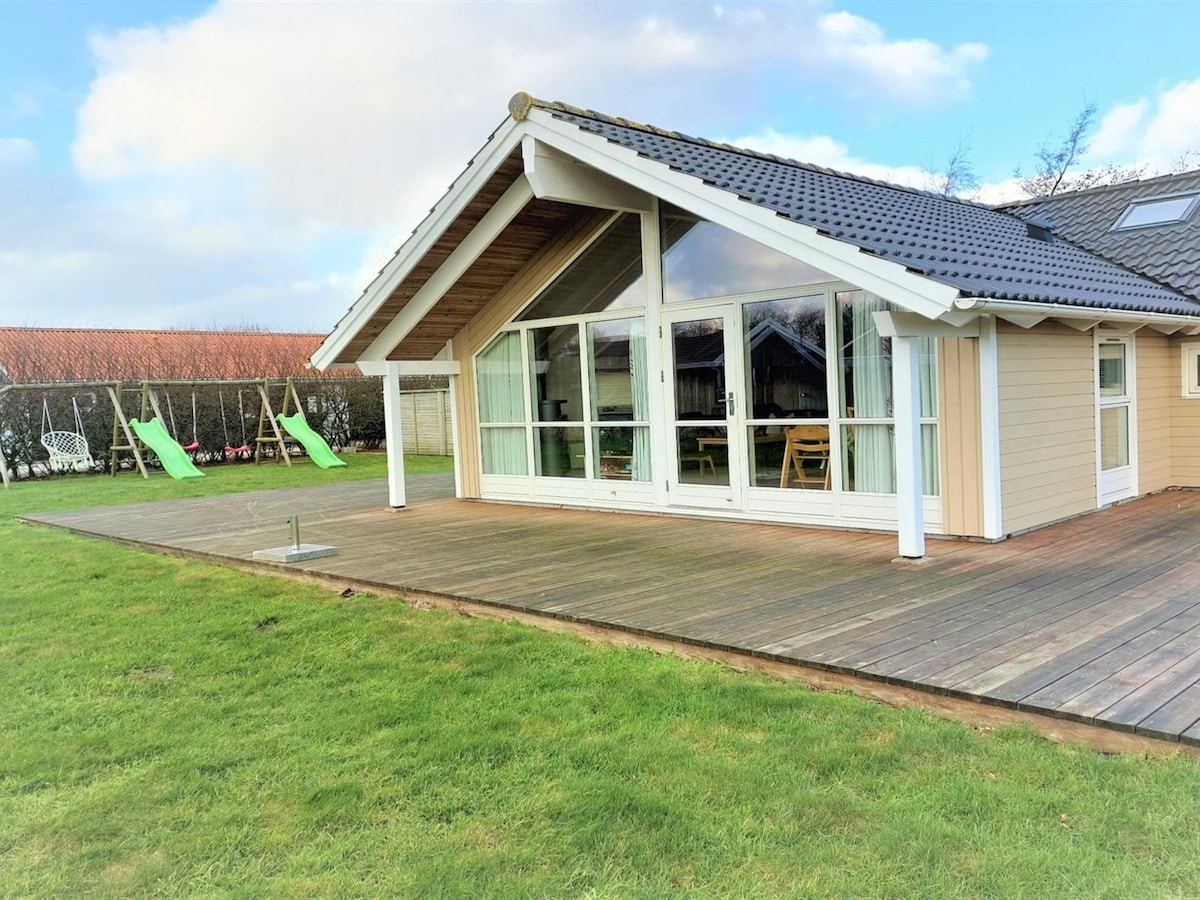 The exterior view of the house displays a sloped roof and large glass doors that open onto a wooden deck. The surrounding area features a grassy lawn and a children's playground with swings in the background.
