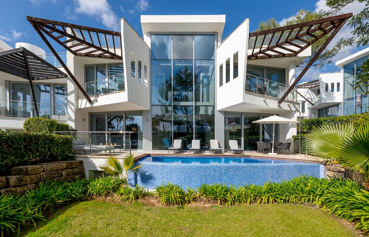 The exterior of the villa is showcased, featuring modern architectural elements and large glass windows. A private swimming pool is surrounded by manicured gardens with lush greenery, while lounge chairs and an umbrella provide a comfortable outdoor seating area.