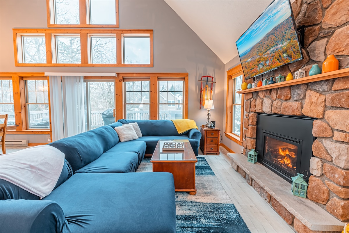A spacious living area features a large, comfortable couch in blue. A stone fireplace is complemented by a wooden coffee table. A wide-screen TV is mounted above the fireplace, and large windows allow natural light to fill the room, enhancing the cozy atmosphere.