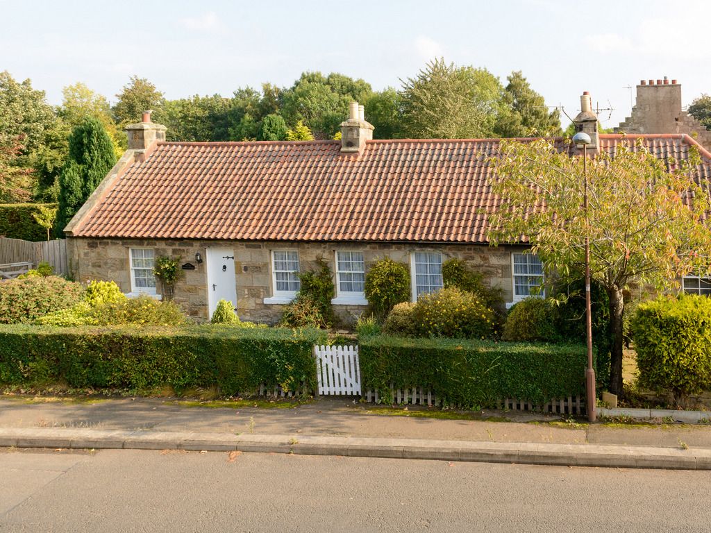 A single-story cottage is visible, characterized by its sandstone exterior and a red-tiled roof. Lush greenery surrounds the property, with well-maintained hedges and a small white picket fence at the front. A quiet street is seen in the foreground, enhancing its tranquil setting.