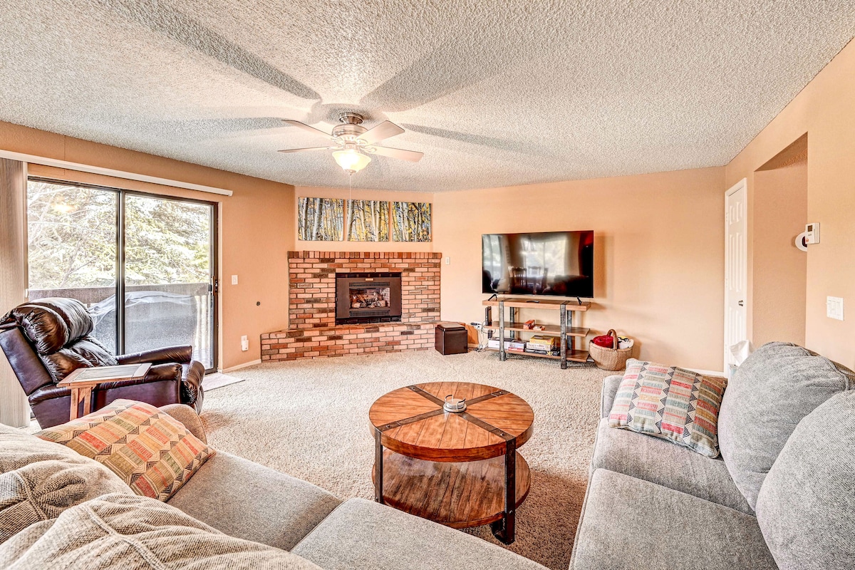 A spacious living area features a large sectional sofa and a wooden coffee table at its center. A flat-screen TV is mounted on the wall opposite a cozy, brick fireplace. Natural light enters through sliding glass doors leading to a balcony.
