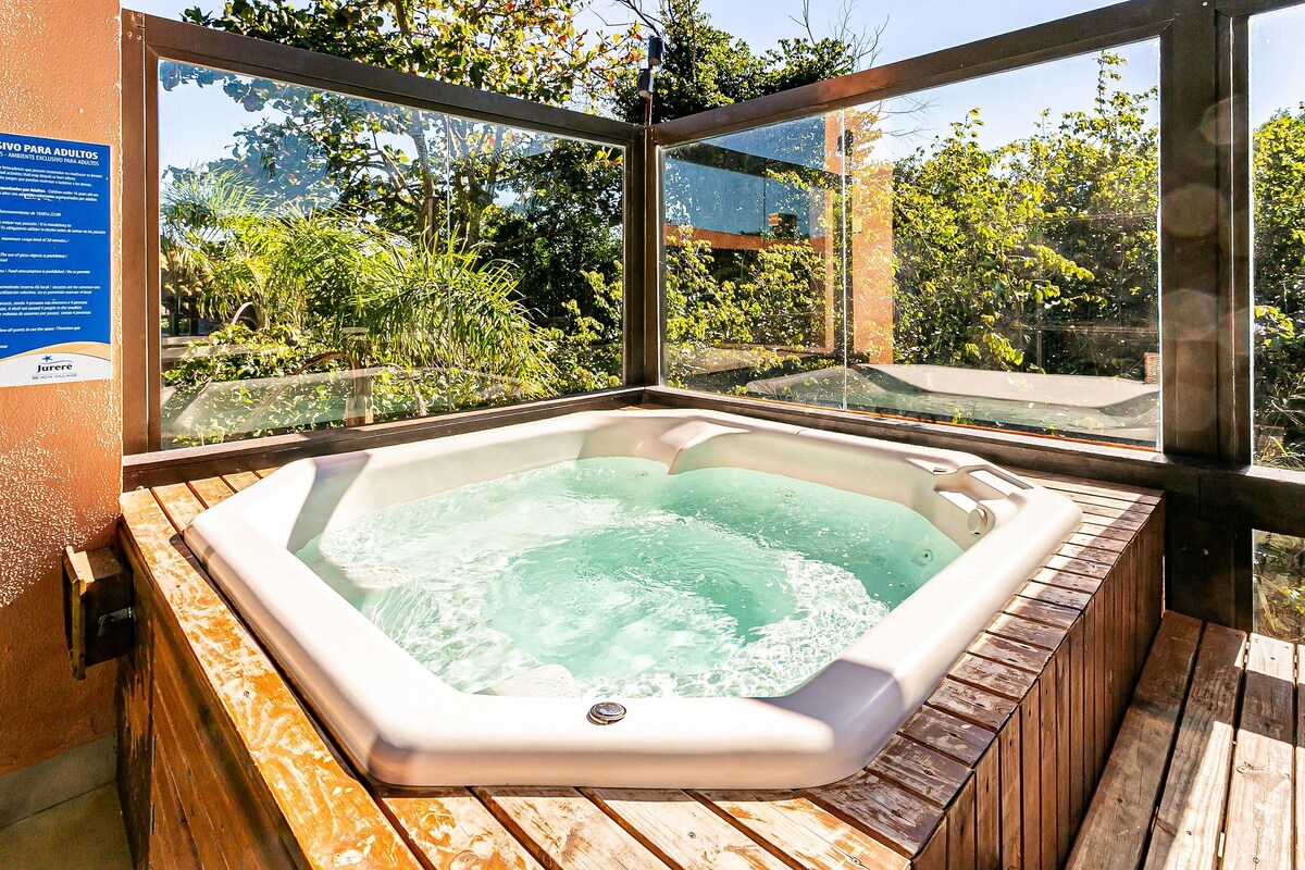 An outdoor jacuzzi is featured within a wooden deck, enclosed by transparent glass panels. Lush greenery surrounds the space, contributing to a serene atmosphere. Sunlight filters through the trees, highlighting the water's surface as gentle bubbles rise.
