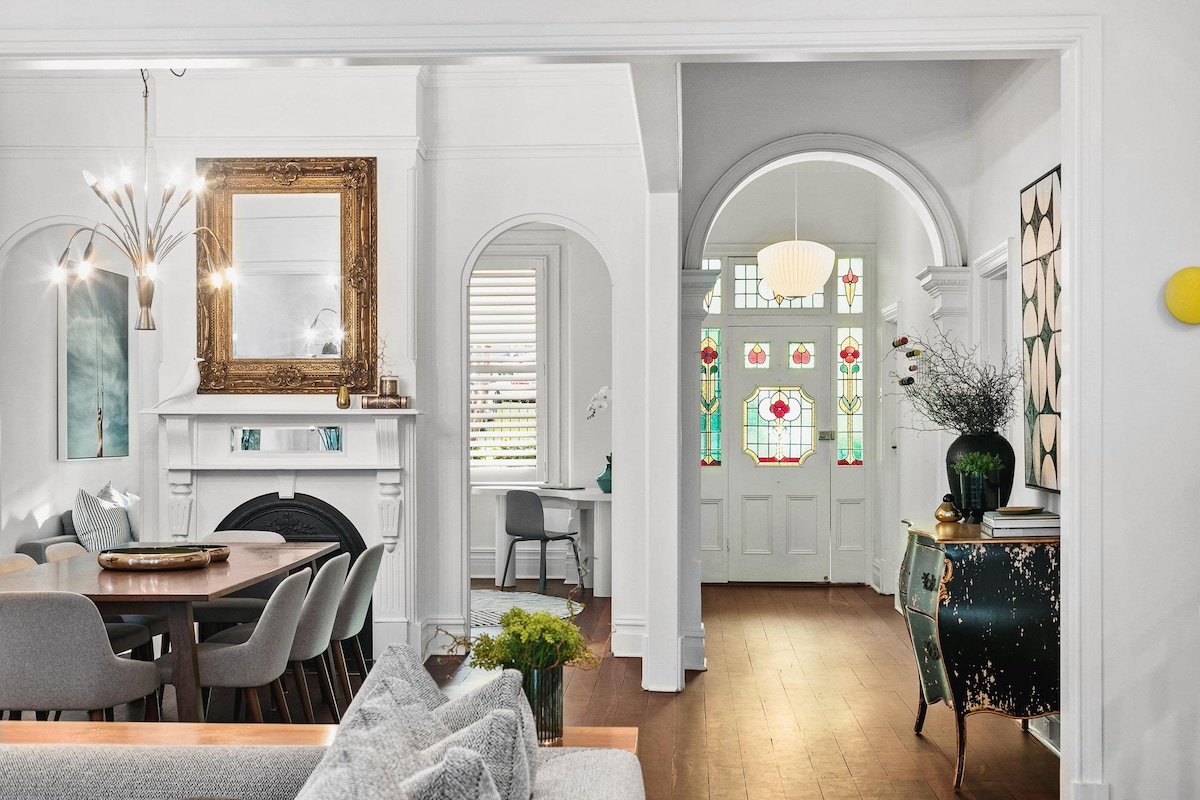 An elegant entrance is highlighted by stained glass windows, welcoming natural light into the home. A spacious dining area features a wooden table surrounded by grey chairs, complemented by a stylish mirror above the fireplace. Decorative elements enhance the inviting atmosphere.