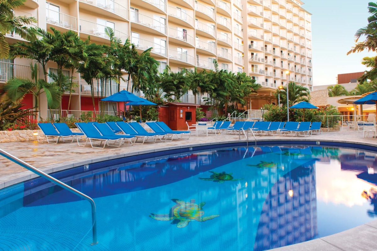 A serene outdoor pool area is surrounded by vibrant tropical foliage and lounge chairs. The water features a turtle design on the pool floor, with blue umbrellas providing shade to the seating. The hotel building stands in the background, showcasing multiple stories.
