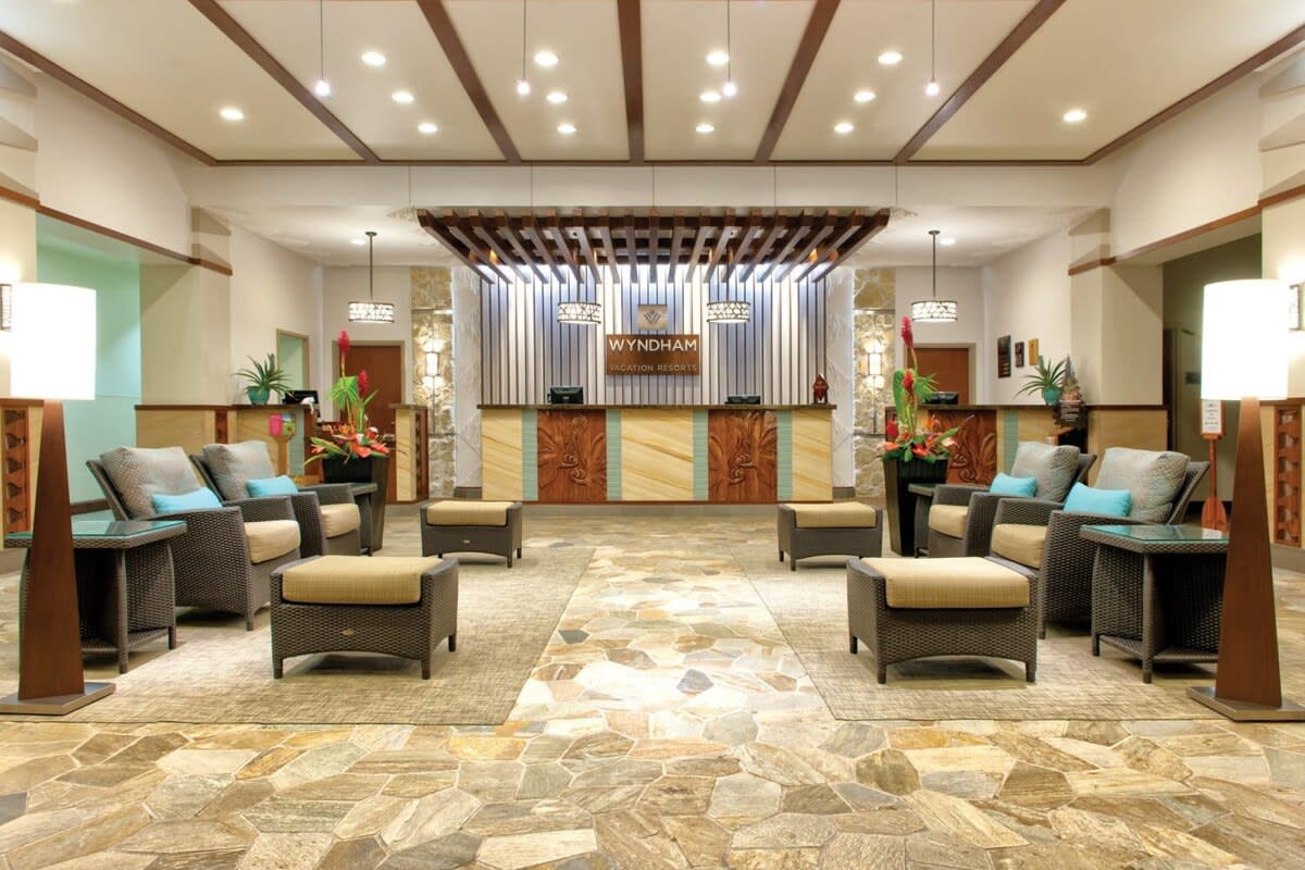 The lobby area features a welcoming reception desk framed by elegant wood beams. Plush seating arrangements with wicker chairs and soft cushions are positioned around a natural stone floor, enhanced by tropical plants and ambient lighting, creating an inviting atmosphere for guests.