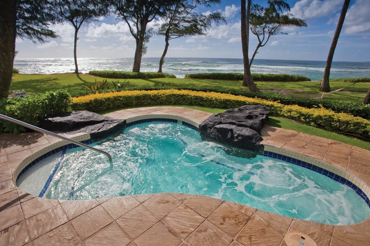 An outdoor hot tub is framed by lush greenery and features natural rock accents. The calming water glistens under sunlight, while the ocean waves can be seen in the background, framed by tall palm trees swaying gently in the breeze.