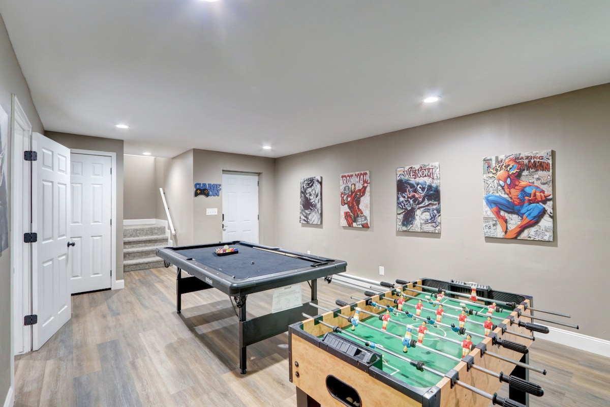A game room is showcased, featuring a pool table and a foosball table. The walls are adorned with comic-inspired artwork. Bright recessed lighting enhances the inviting atmosphere, while a staircase is visible in the background, leading to other areas of the chalet.