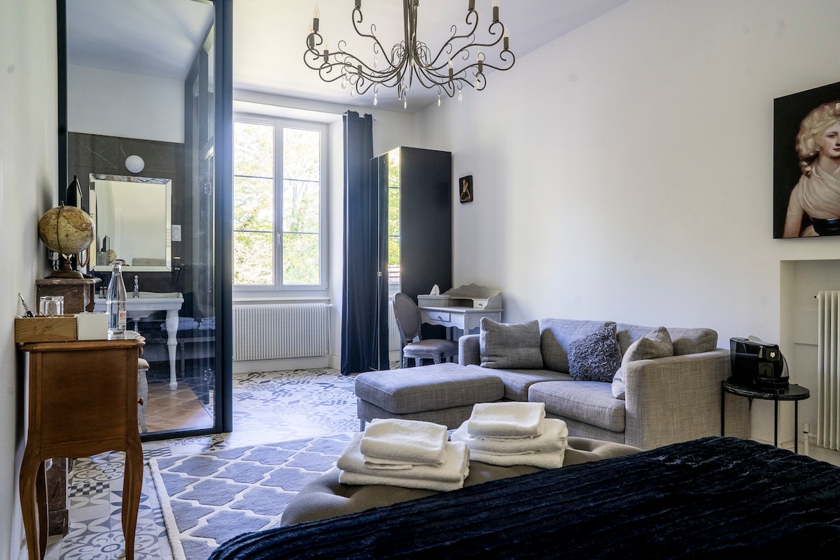 The spacious room features a comfortable seating area with a light gray sofa and decorative cushions. A large window allows natural light to fill the space, which includes a stylish chandelier and a work desk. A glass partition reveals the adjoining bathroom, showcasing elegant decor.