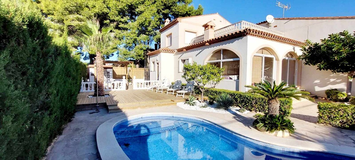 A serene outdoor area features a private swimming pool surrounded by a well-maintained garden with palm trees and hedges. A wooden deck is visible, furnished with lounge chairs, and a terrace with arches offers additional seating for relaxation.