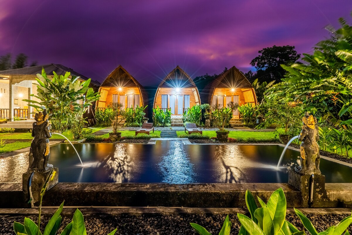 Four charming bungalows are framed by lush greenery, facing a tranquil pond with gentle water fountains. Soft lighting enhances the evening ambiance, while decorative statues add cultural elements to the garden. The scene offers a serene retreat among the natural surroundings.