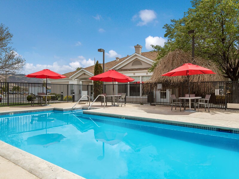 An outdoor swimming pool is surrounded by a safety fence and features bright red umbrellas over lounge seating. The pool area is enhanced by a clear blue sky and well-maintained landscaping in the background.
