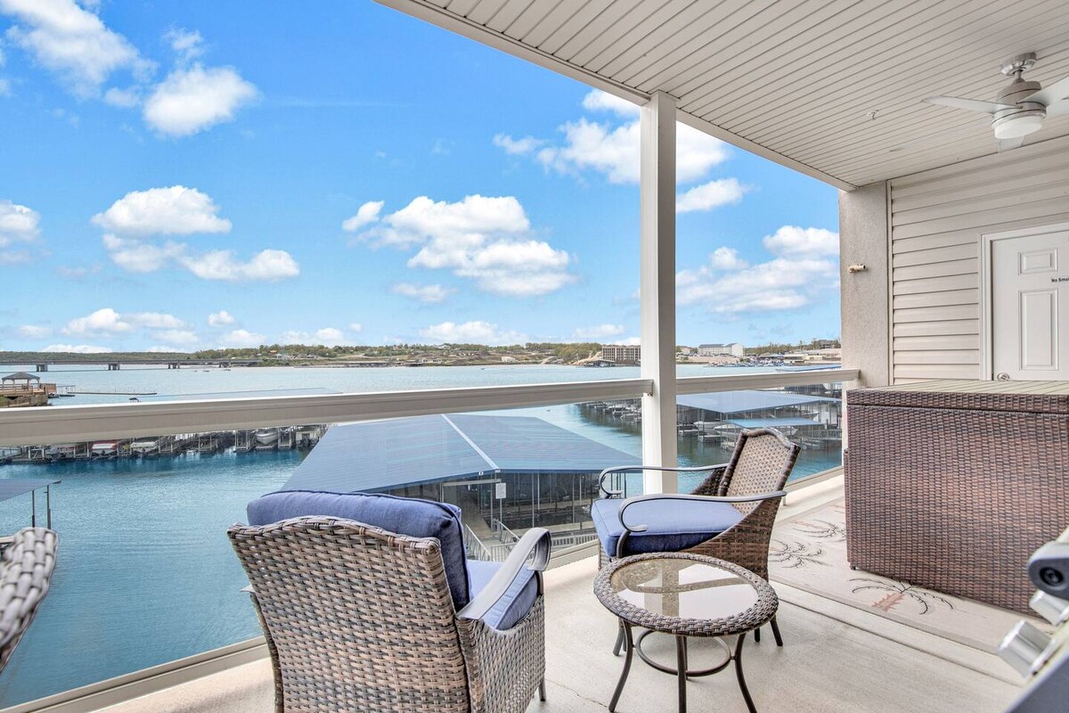 A spacious outdoor deck overlooks the sparkling water, featuring comfortable wicker seating and a glass-top table. Panoramic views of the lake and surrounding area are visible, creating a serene environment. A ceiling fan is positioned above, enhancing the comfort of the space.