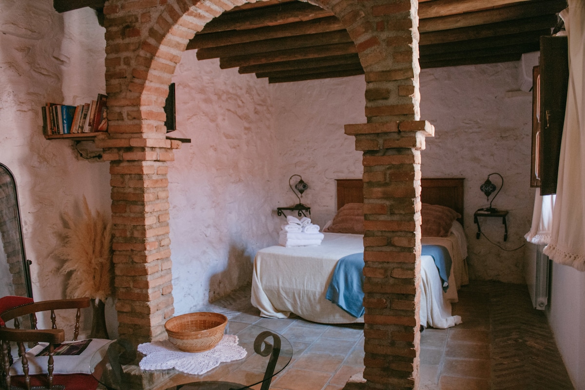 A cozy room features a spacious bed, flanked by two reading lamps. Exposed wooden beams highlight the rustic ceiling, while a decorative arch separates the sleeping area from a small dining space with a round table and chairs. A warm light filters through a nearby window.