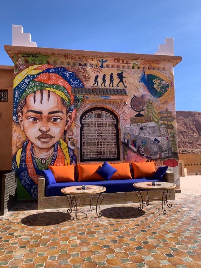 A vibrant mural adorns the wall behind a comfortable seating area, featuring a long blue couch with orange cushions. Two round tables sit in front of the couch, placed on a mosaic tiled floor. The setting captures a blend of cultural art and relaxation.