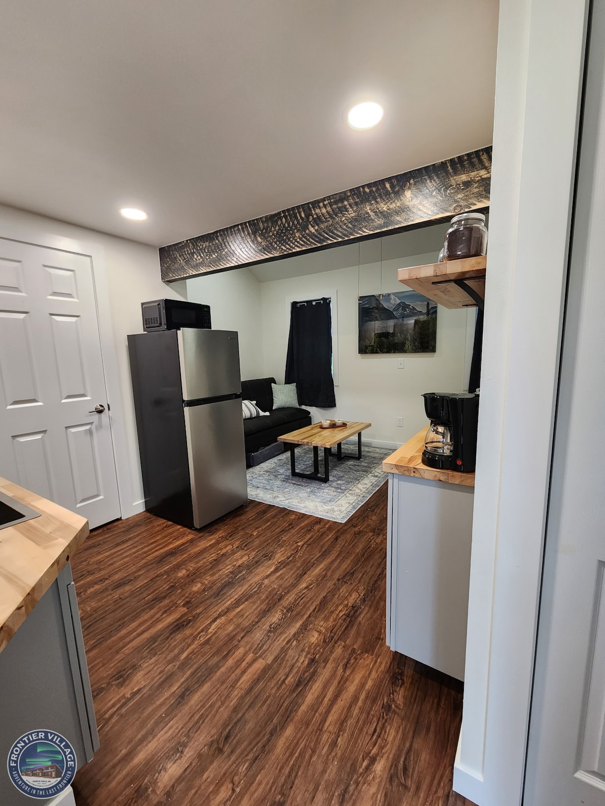 A modern kitchen area features stainless steel appliances, including a refrigerator and coffee maker. A wooden countertop offers space for meal preparation, while an open living area is visible, showcasing a dark sofa and a coffee table. Bright lighting enhances the welcoming atmosphere.