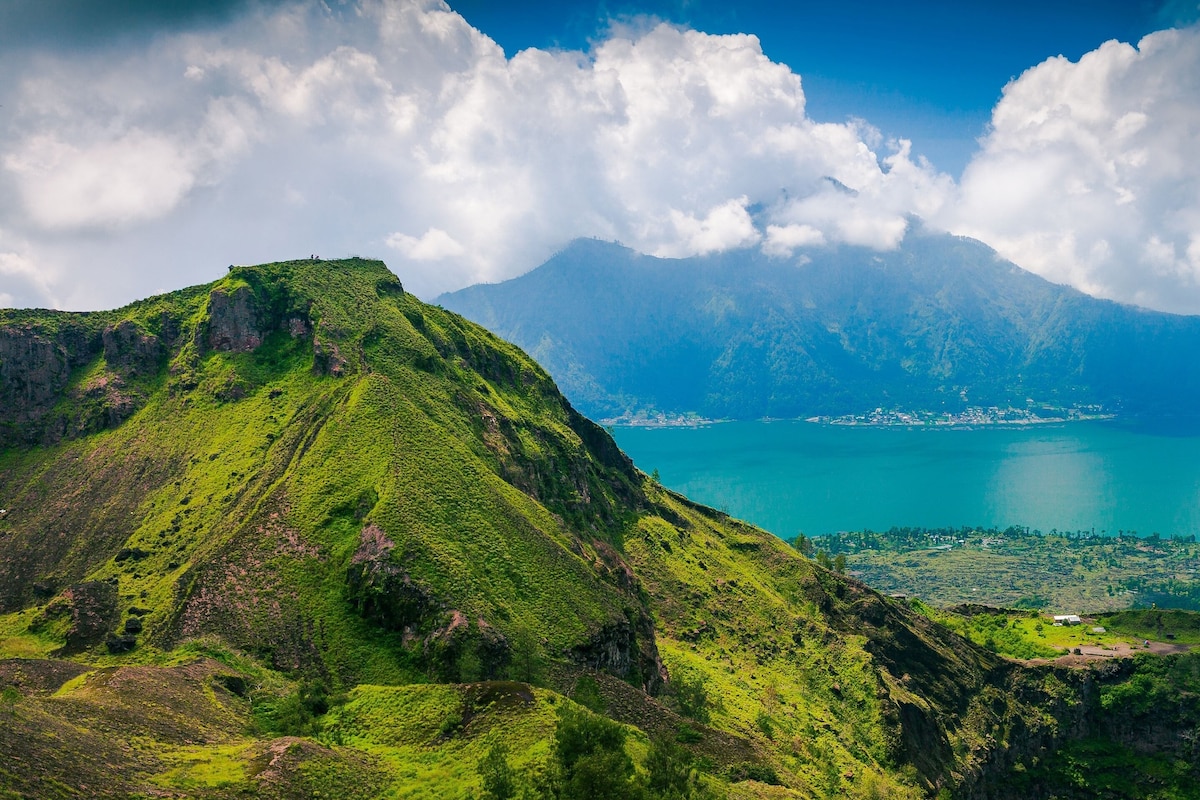 Lush green hillsides are viewed overlooking Lake Batur, framed by dramatic mountain silhouettes. The vibrant landscape blends with a bright blue sky dotted with fluffy white clouds. This serene setting captures the natural beauty of the area surrounding the bungalows.