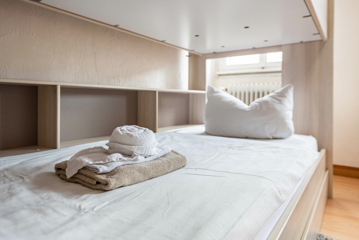 A neatly arranged sleeping area features a spacious bed with a white pillow and a soft blanket. Freshly laundered towels are stacked nearby, emphasizing the cleanliness of the space. Natural light enters through a window, illuminating the calm ambiance.
