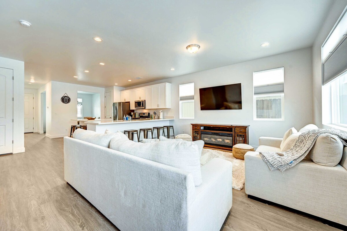 An open-concept living space is presented with light-colored walls and a bright, airy feel. A spacious sectional sofa invites relaxation, while a large smart TV is mounted on the wall. The kitchen area features a breakfast bar with stools and modern appliances.