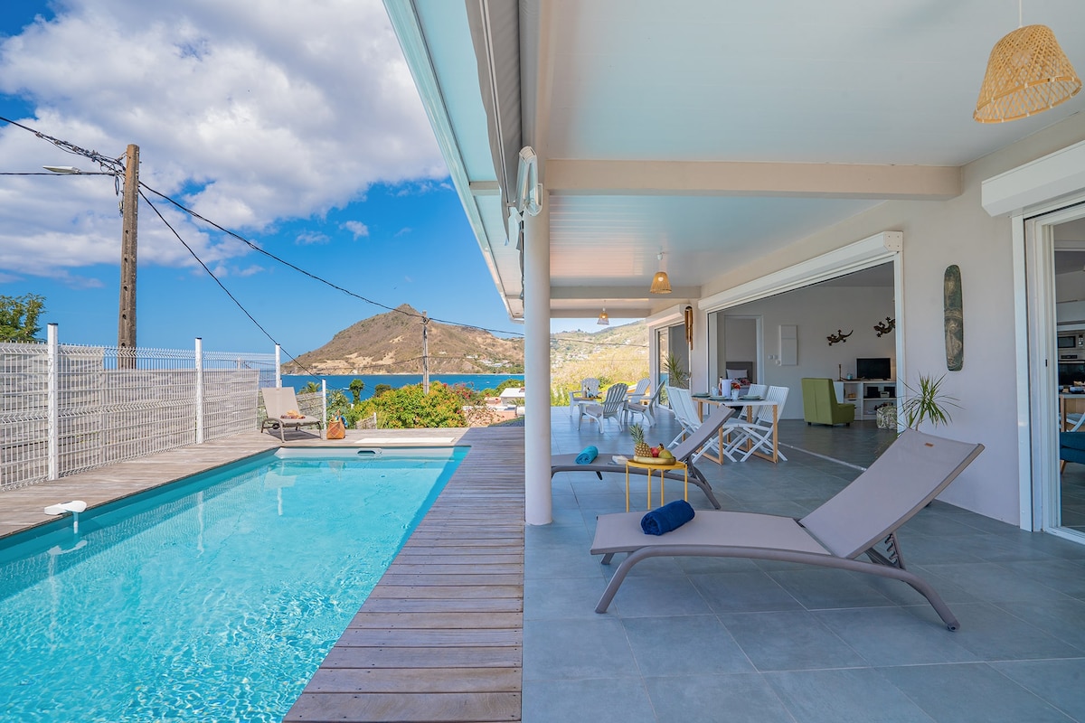 A clear view of the serene outdoor space is presented, featuring a refreshing pool beside a spacious patio. Sun loungers are positioned near the pool, with coastal scenery visible in the background, including lush hills and the sea under a partly cloudy sky.