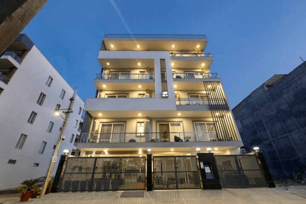 A modern, multi-story building is illuminated by exterior lights, revealing spacious balconies on each floor. The facade features a combination of sleek lines and a decorative gate at the entrance. The surrounding area showcases a mix of urban architecture.