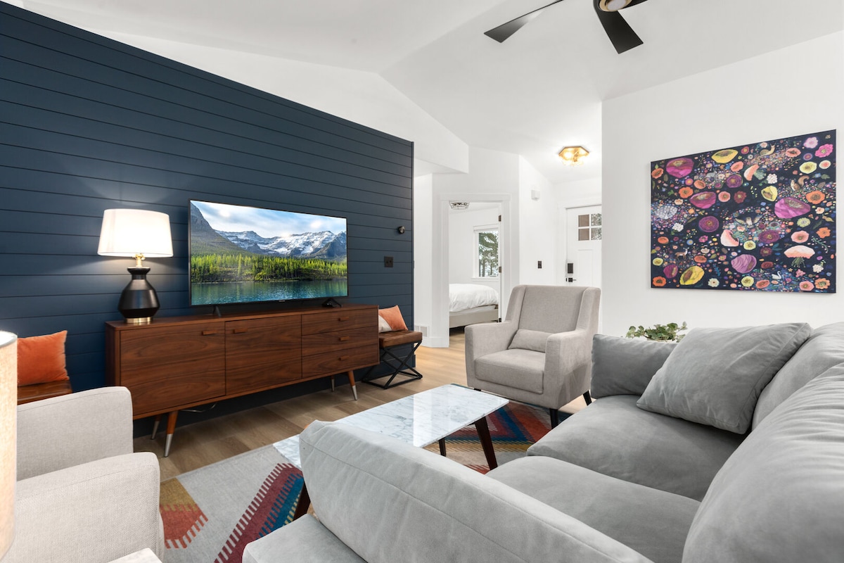 A modern living space showcases a large flat-screen television mounted on a blue accent wall. Comfortable grey couches are arranged around a glass coffee table, while a colorful wall art piece adds visual interest. Natural light filters in from nearby windows, enhancing the room's inviting ambiance.