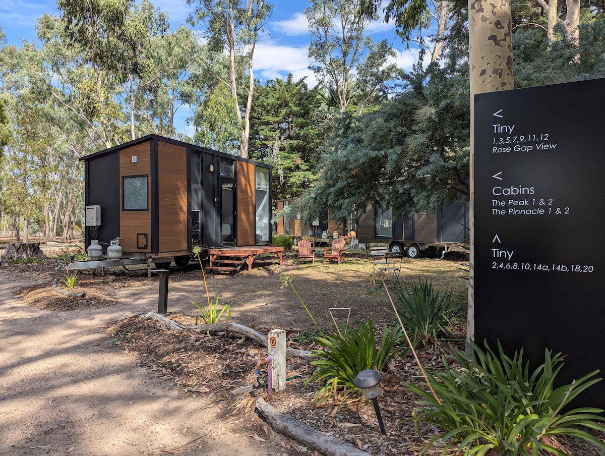 A tiny house is nestled among trees, showcasing a modern exterior with dark panels and large windows. A wooden deck extends from the entrance, accompanied by outdoor seating. Nearby, additional cabins are visible, framed by lush greenery and marked by directional signage.