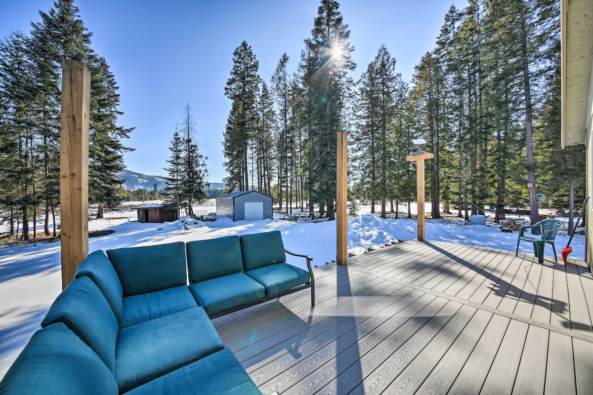 A deck with lounge seating is presented, surrounded by tall trees and a snowy landscape. Sunlight filters through the branches, creating a bright and inviting atmosphere. A distant shed is visible, complementing the serene setting.