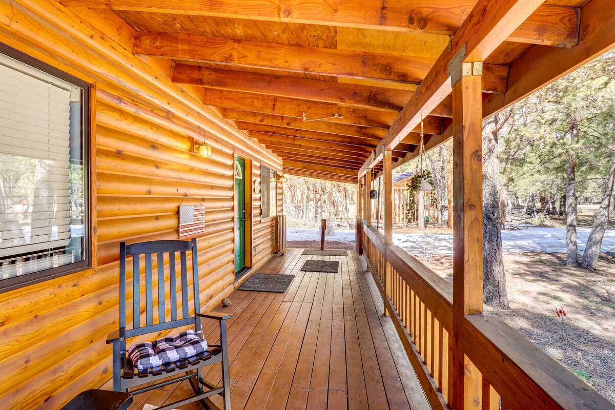 A wooden porch showcases a spacious layout with natural wood flooring and ceiling beams. A rocking chair is positioned on one side, offering a place to relax. Views of the surrounding forest and pathway can be seen through the open space of the railing.