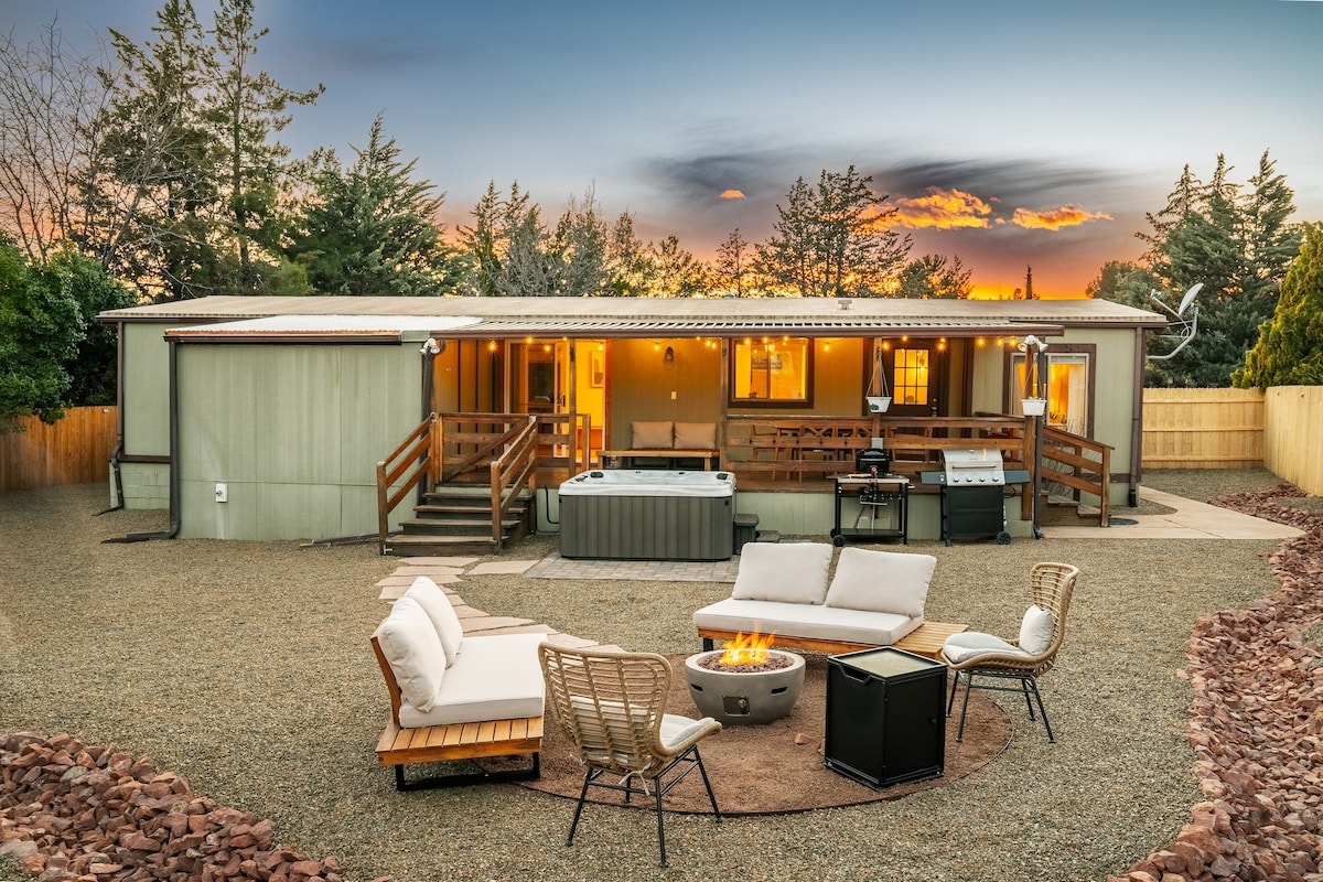An outdoor space features a cozy fire pit surrounded by comfortable seating, with a luxurious hot tub nearby. The backdrop includes a spacious dwelling with warm lighting, complemented by a well-equipped outdoor kitchen. Trees and a sunset sky enhance the serene atmosphere.