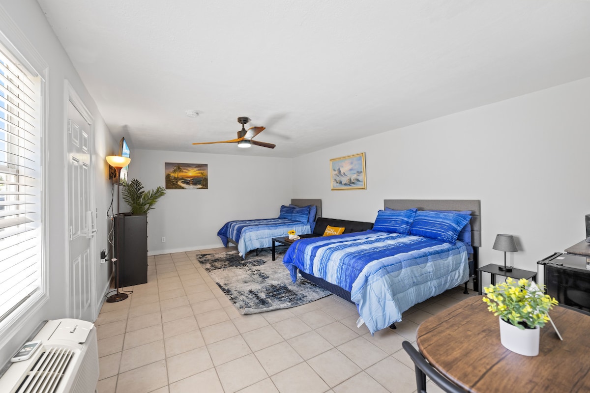 The airy studio features two spacious beds dressed in blue linens, positioned on a tiled floor. A ceiling fan provides airflow. A small table accompanies a chair, while a potted plant and wall art add warmth to the room. Bright natural light enters through the window.