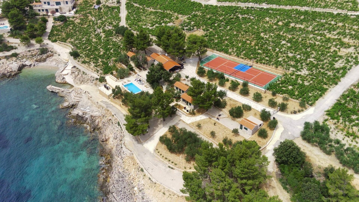 An aerial view showcases a private resort by the sea, surrounded by lush vineyards and greenery. The image highlights a swimming pool, tennis court, and well-placed paths interspersed among the buildings, all set against the tranquil blue waters of the Adriatic.