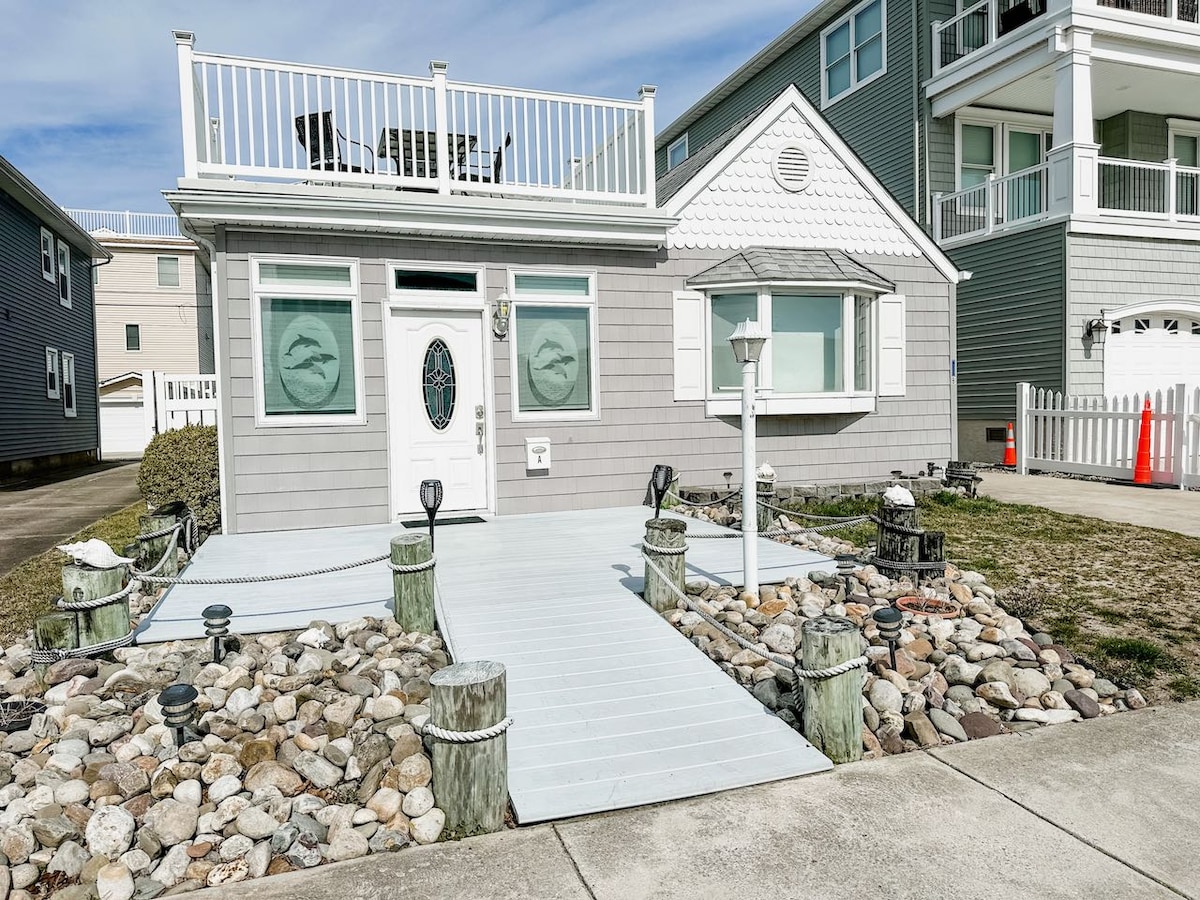 The exterior of a charming gray house is depicted, featuring a covered front porch and decorative stone landscaping. The entrance is framed by a welcoming pathway, with two unique lanterns and a light fixture positioned near the door. A spacious roof deck is visible above.