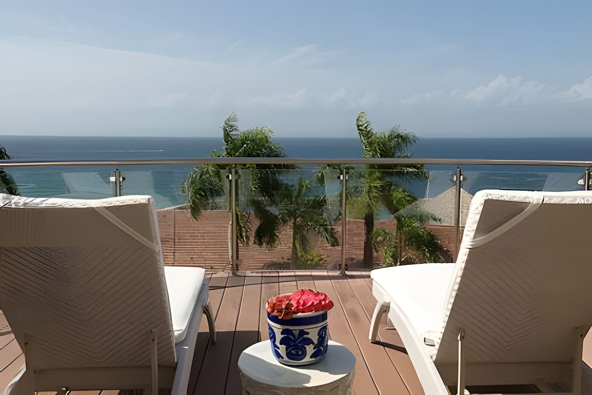 Two white lounge chairs are positioned on a wooden terrace overlooking the ocean. A small round table with a decorative bowl of pink flowers sits between the chairs. Lush greenery and clear blue skies enhance the serene atmosphere.