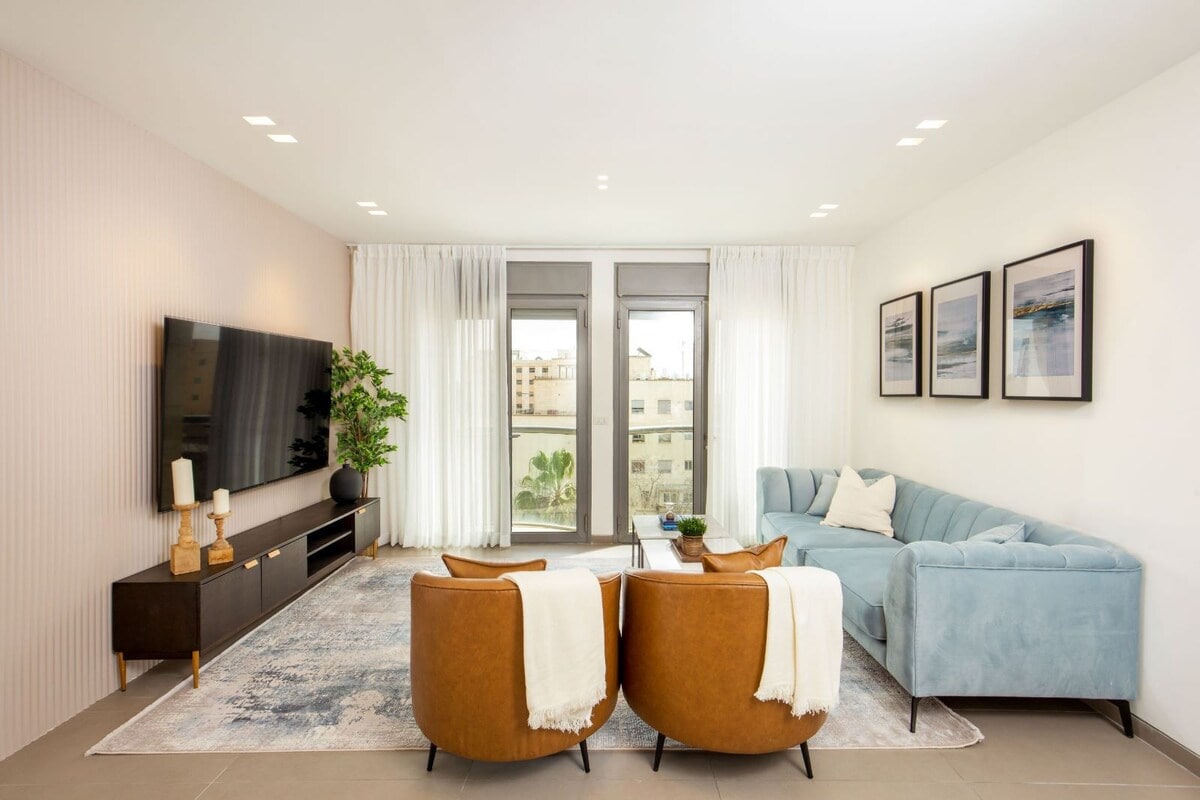 A bright living area features a comfortable light blue sofa and two tan armchairs, complemented by a decorative rug. Large windows invite natural light, with sheer curtains softly framing the view. A modern TV is mounted on the wall, and framed artwork adds an elegant touch.