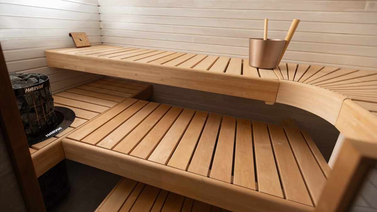 The sauna features wooden benches arranged in a tiered layout, providing ample seating. A wooden bucket and ladle are positioned nearby. The walls are clad in light-colored wooden panels, enhancing the serene atmosphere. The floor is composed of dark tiles, contrasting with the lighter wood.